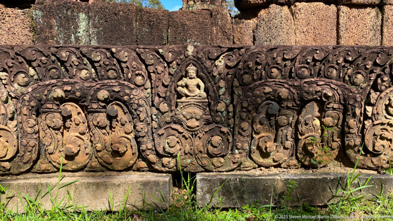 Ground-level carved sandstone blocks with seated figures, monster masks, and vegetal scrollwork at Banteay Srei.