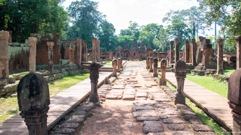 Long stone causeway lined with short posts, wooden walkways, and temple ruins.
