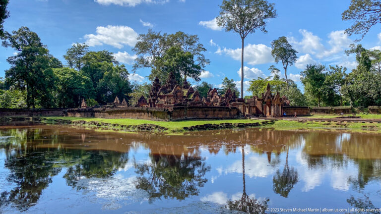 Wide view of Banteay Srei reflected in a pond beneath trees and blue sky.