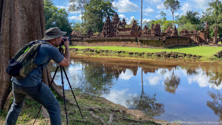 Photographer using a tripod beside a pond with Banteay Srei reflected across the water.
