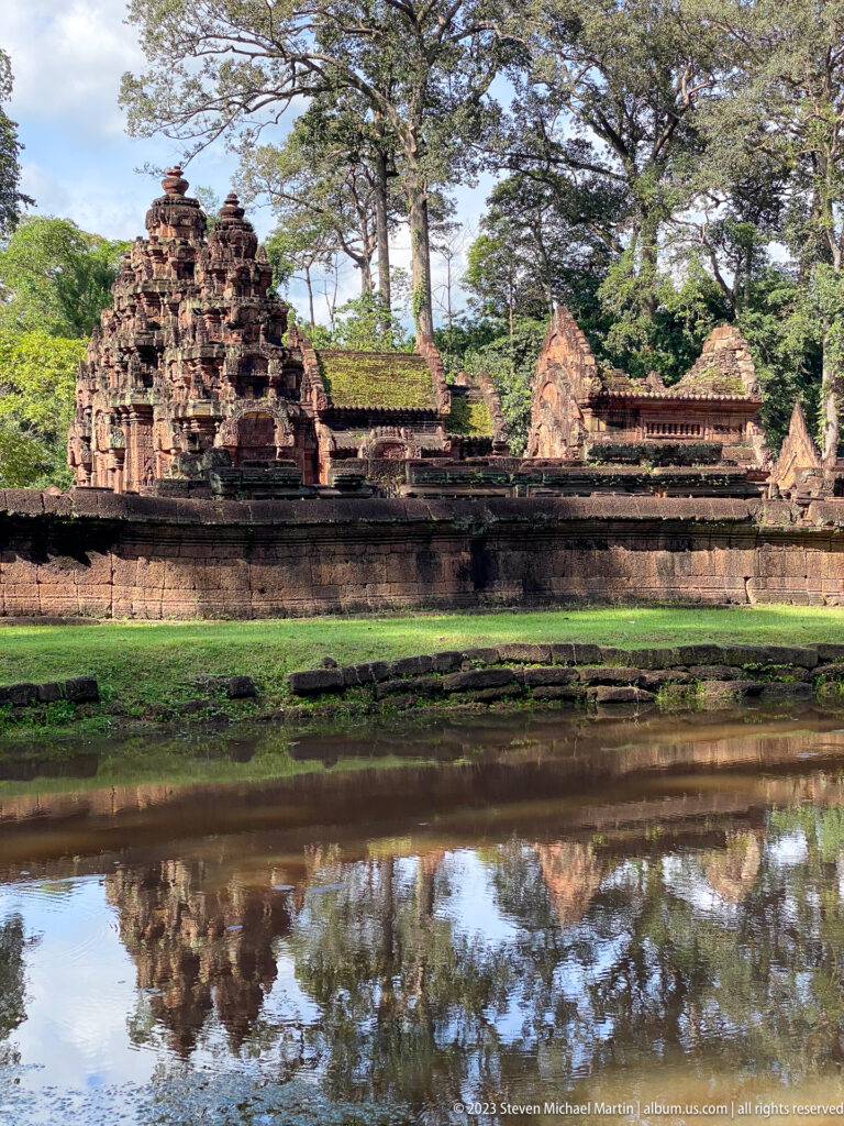 Side view of Banteay Srei enclosure walls and small towers reflected across a pond.