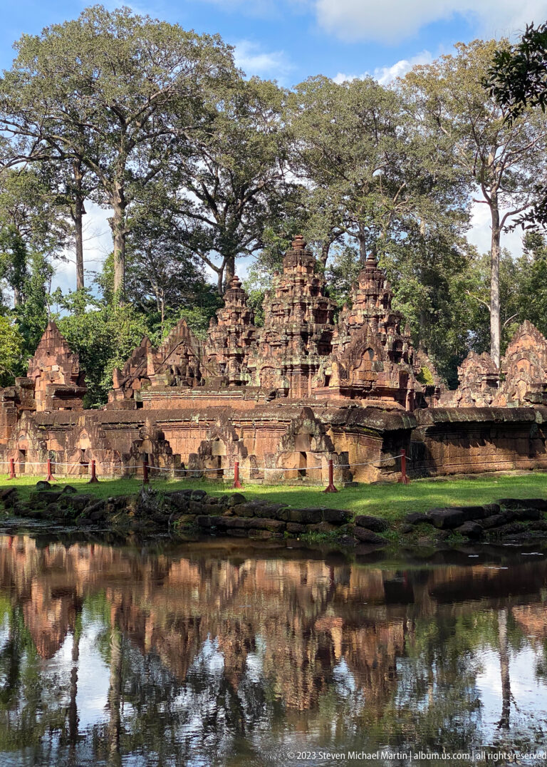 Wide panoramic view of Banteay Srei towers and trees reflected in a calm pond.