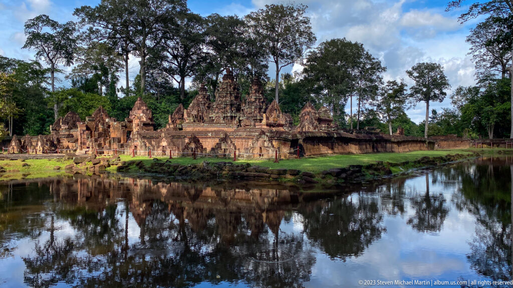 Centered wide view of Banteay Srei and surrounding trees reflected in a still pond.