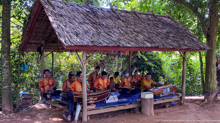 Group of Cambodian musicians performing traditional instruments under a thatched shelter near Banteay Srei.