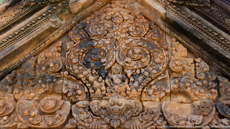 Triangular sandstone pediment with a seated deity, curling ornament, and a large monster face below.