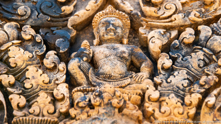 Close-up of a crowned seated figure carved in sandstone and surrounded by curling foliage.