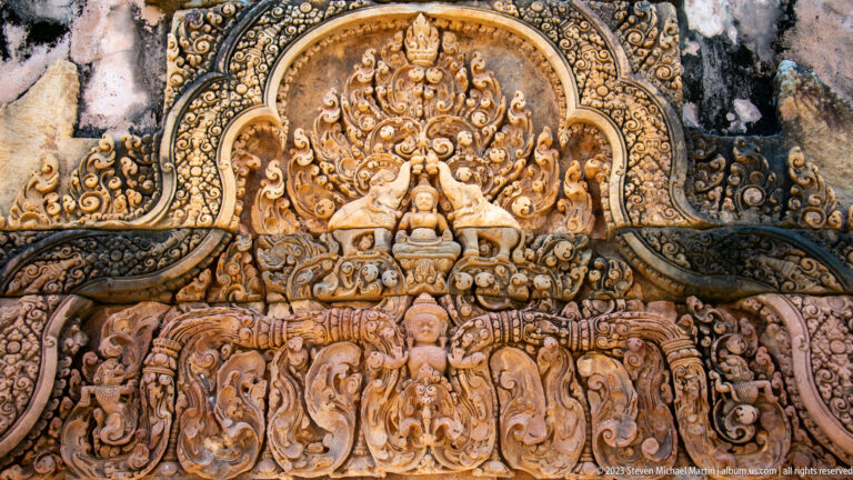 Arched sandstone relief with a seated central figure, two guardian animals, and smaller figures below.