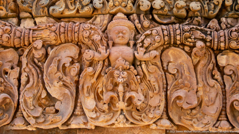 Standing female figure with raised hands and tall headdress surrounded by symmetrical sandstone scrollwork.