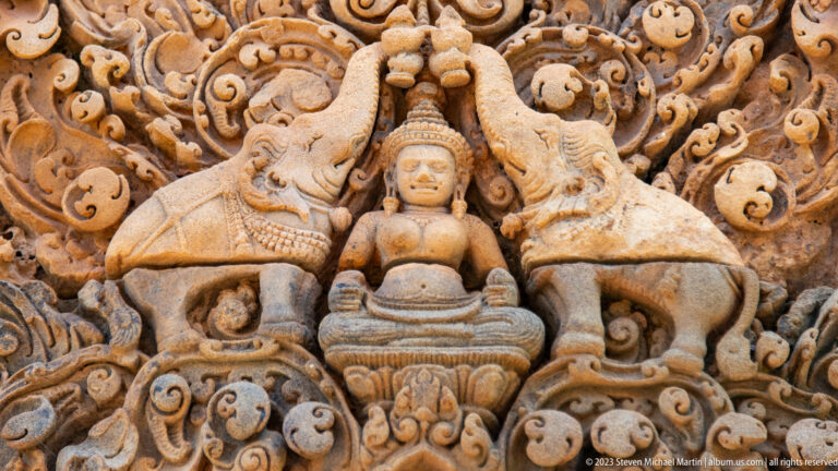 Seated sandstone figure framed by two inward-facing animals and dense curling ornament.