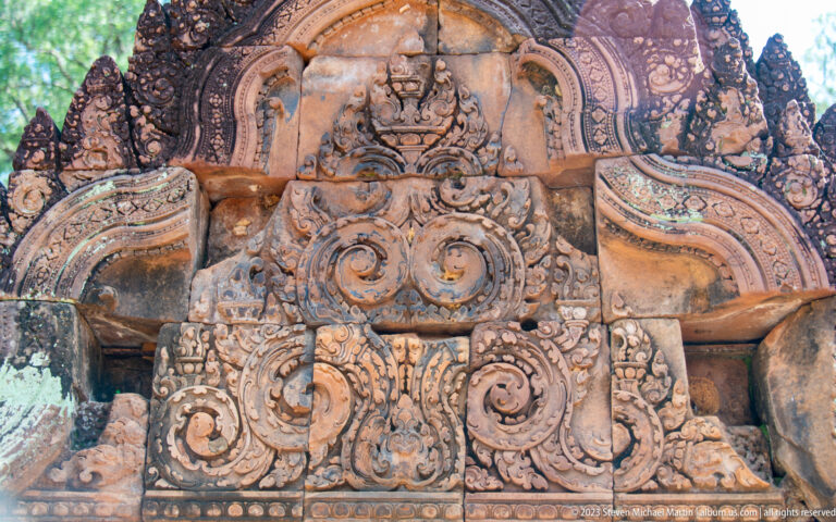 Tall weathered sandstone pediment with large scrolls, damaged figures, and repaired sections.