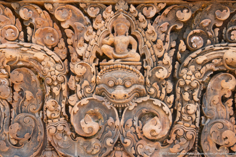 Seated figure carved above a large round-eyed monster face amid curling sandstone foliage.