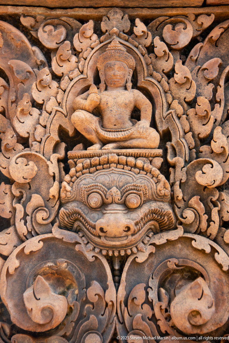 Extreme close-up of a large carved monster face beneath the knees of a seated figure.