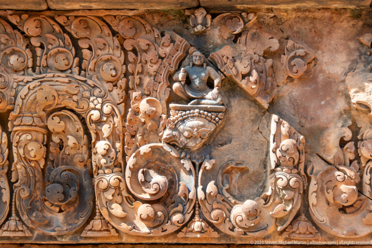 Damaged sandstone relief with a small seated figure, dark mask-like form, and large area of stone loss.