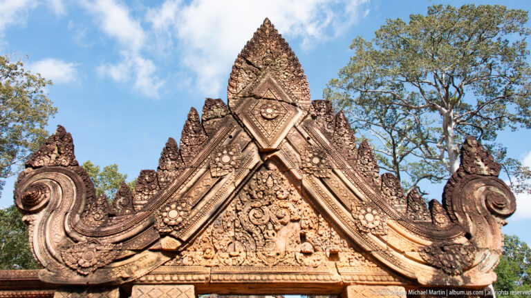Complete pointed sandstone gateway gable with curved sides, floral medallions, and blue sky behind.