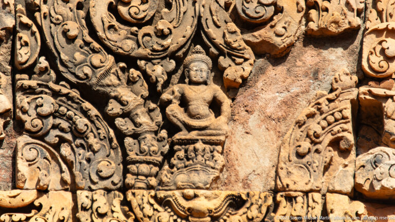 Close-up of a damaged sandstone relief with a seated figure, mask motif, and missing stone.