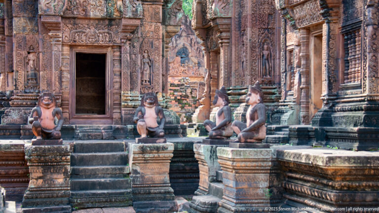 Four seated monkey-like guardian statues flank steps leading to an intricately carved pink-sandstone temple doorway.