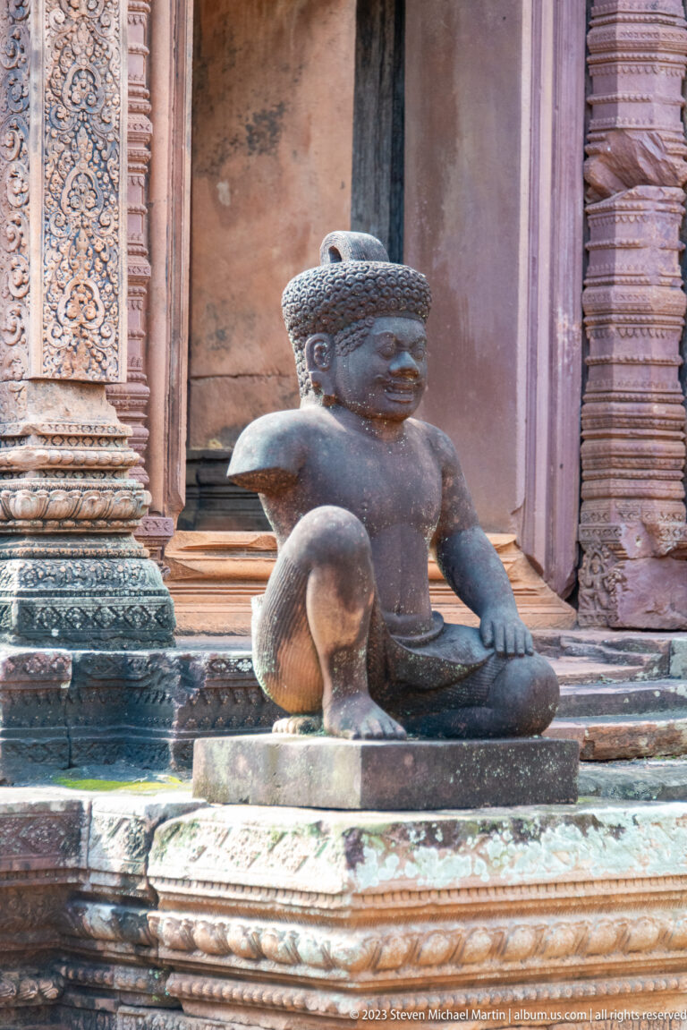 Close view of a seated monkey-like guardian with one knee raised beside a carved sandstone doorway.