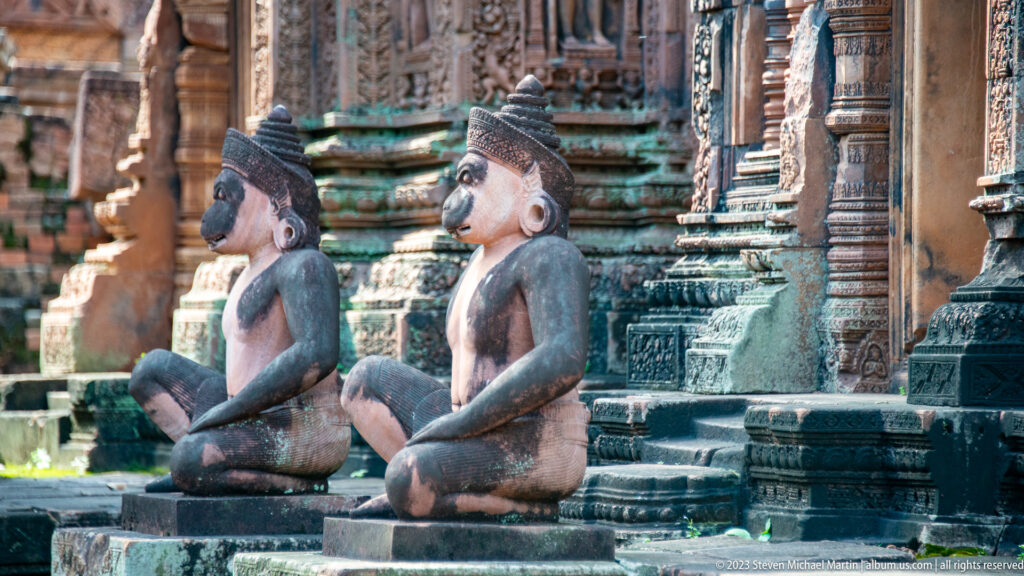 Two seated monkey-like guardian statues shown in profile beside carved sandstone temple walls and platforms.