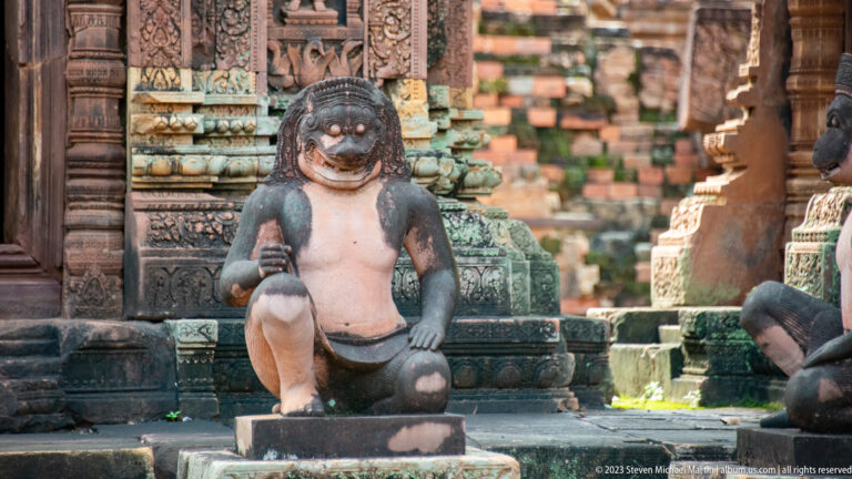 Front-facing monkey-like guardian kneels with one knee raised against an ornate carved sandstone background.
