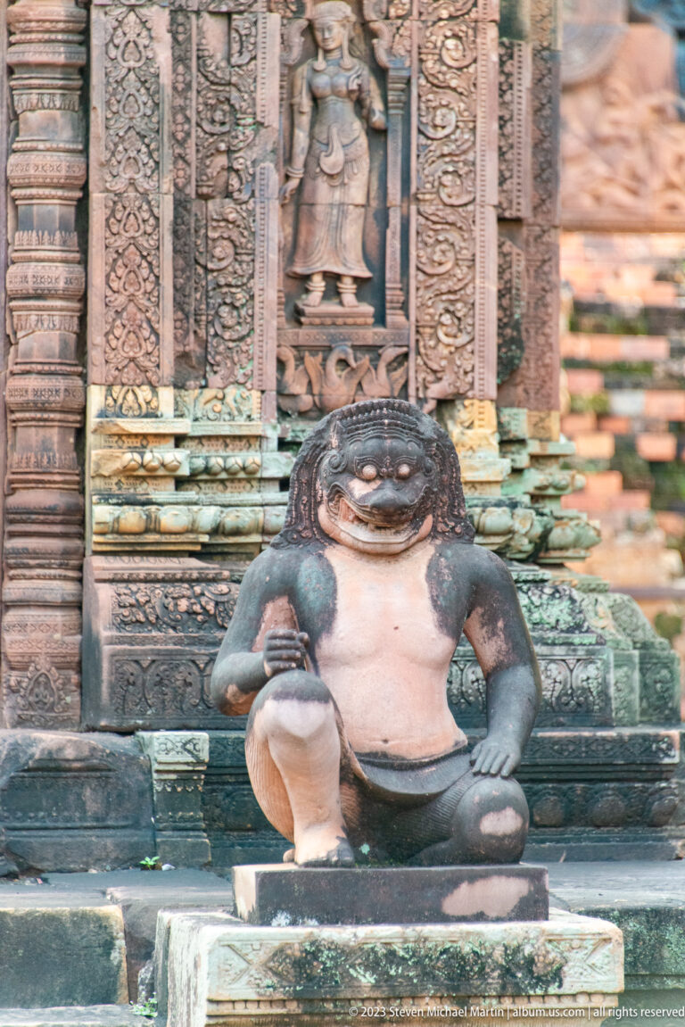 Seated monkey-like guardian with a pale torso and dark limbs framed by ornate sandstone carvings.