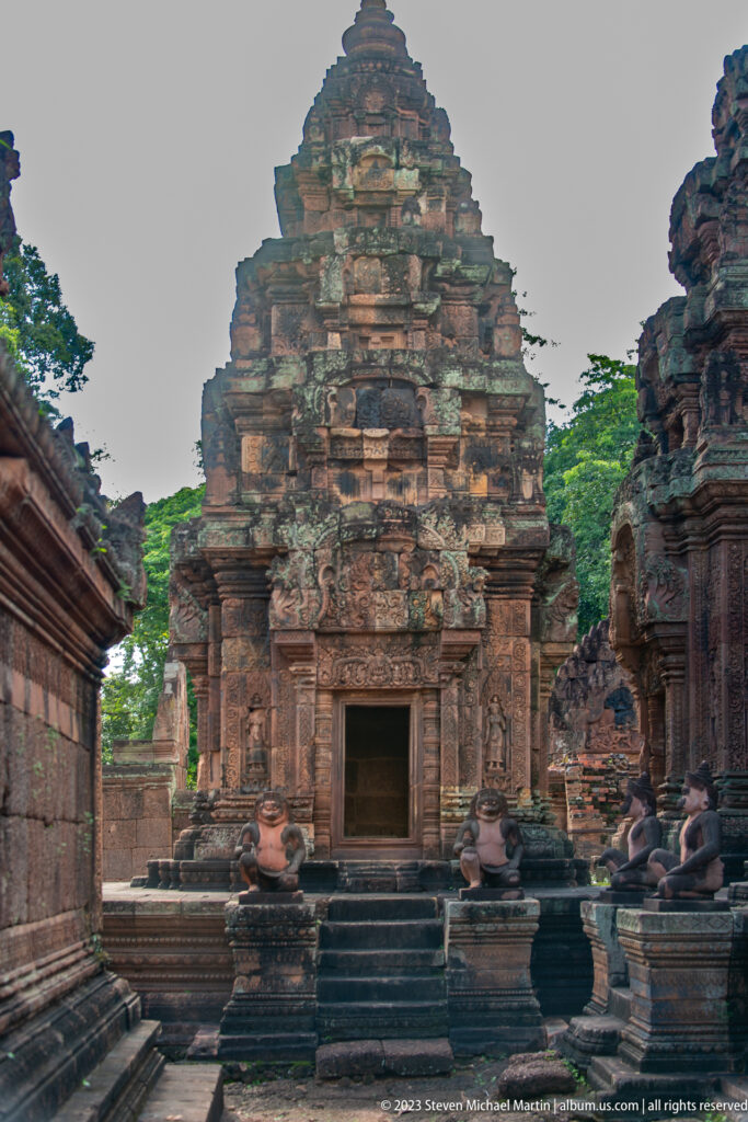 Frontal view of a tiered pink-sandstone sanctuary with steps, a dark doorway, and seated guardians on both sides.