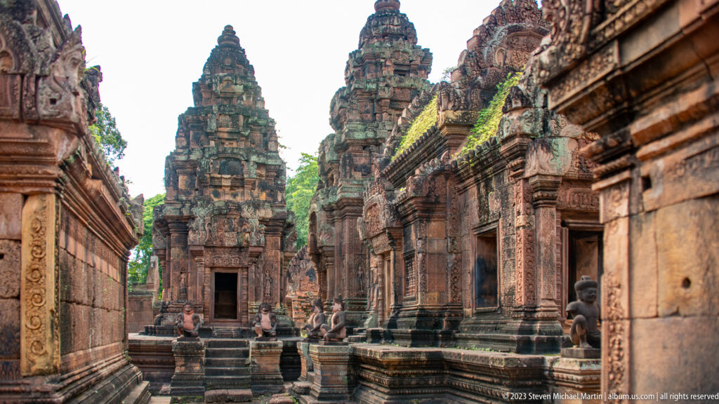 Narrow pink-sandstone temple court leading toward a tall carved sanctuary tower, with seated guardian statues along the path.