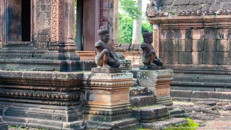 Two dark seated guardian statues on carved pedestals beside an ornate pink-sandstone temple doorway.