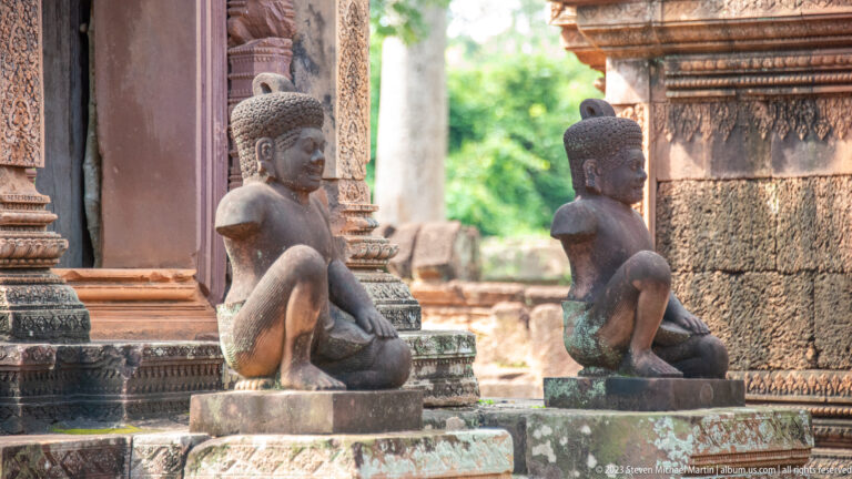 Close side view of two weathered seated guardian statues with rounded headdresses beside carved sandstone architecture.