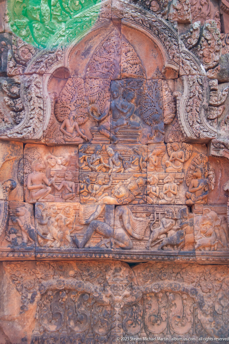 Reddish sandstone pediment carved with a central seated deity, surrounding figures, and dense floral ornament.