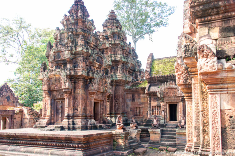 Three carved pink-sandstone sanctuary towers on a raised platform, with guardian statues and an ornate doorway in the foreground.