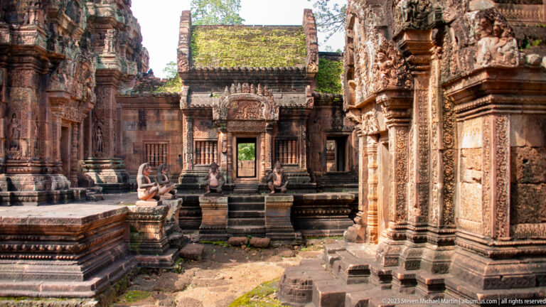 Sunlit temple courtyard leading to a central doorway, with seated guardian statues and richly carved sandstone walls on both sides.