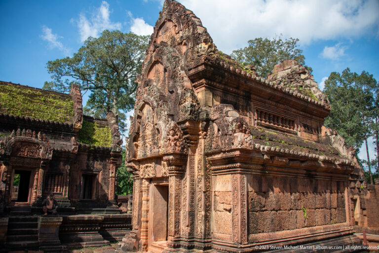 Oblique view of a long carved pink-sandstone temple building with a tall pediment and moss-covered roofline.