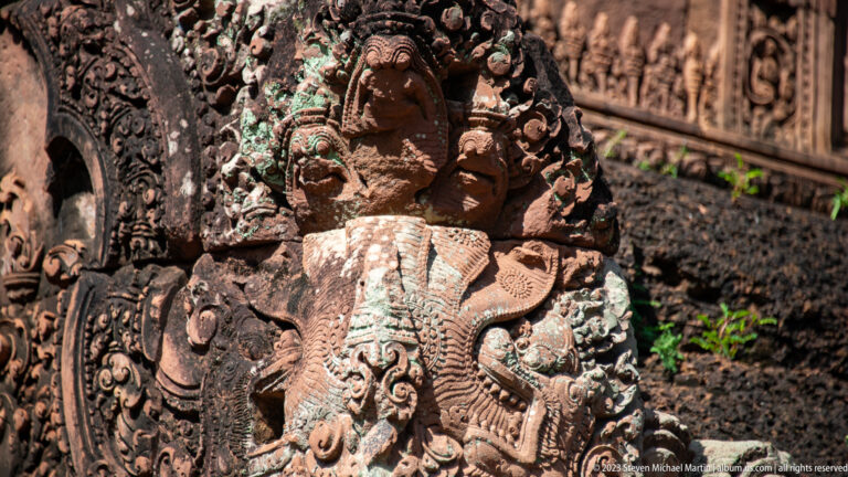 Close oblique view of a carved mythical face surrounded by dense sandstone foliage, small figures, and green lichen.
