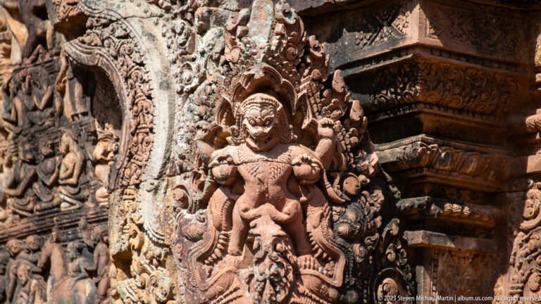 Standing crowned figure in high relief beside an ornate sandstone doorway, wearing a tall headdress and elaborate jewelry.