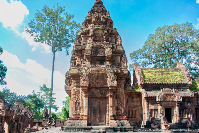 Tall tiered pink-sandstone sanctuary tower with a central doorway, flanked by lower carved temple buildings and trees.