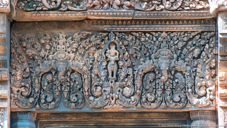 Rectangular sandstone relief with a central standing figure, smaller attendants, and dense symmetrical floral scrollwork.