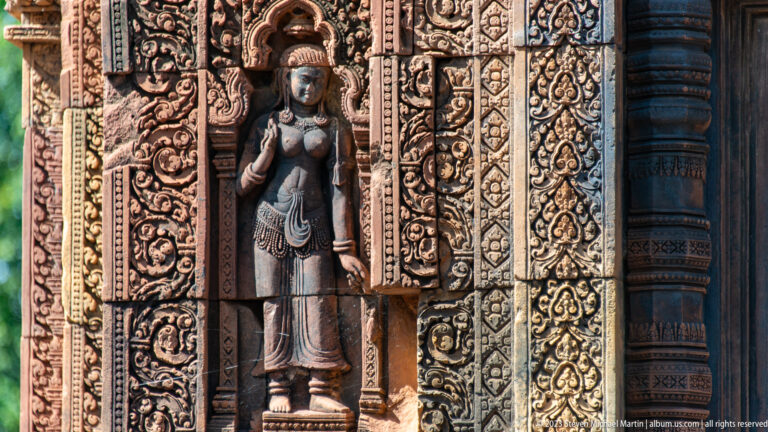 Standing female figure in a carved sandstone niche, wearing a tall headdress, jewelry, and a long garment.