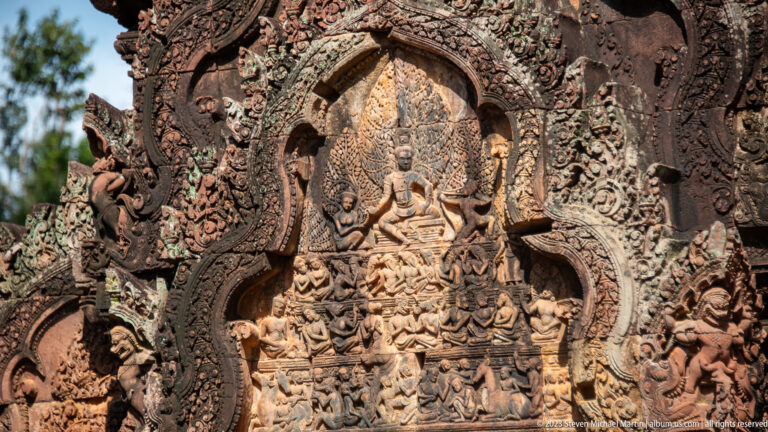 Pointed sandstone pediment with a seated multi-armed deity, smaller figures, and dense carved ornament.