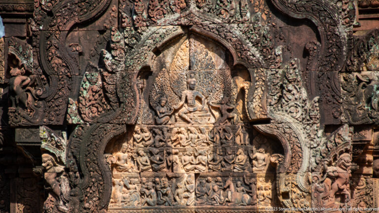 Pointed sandstone pediment with a seated multi-armed deity above rows of smaller carved figures.