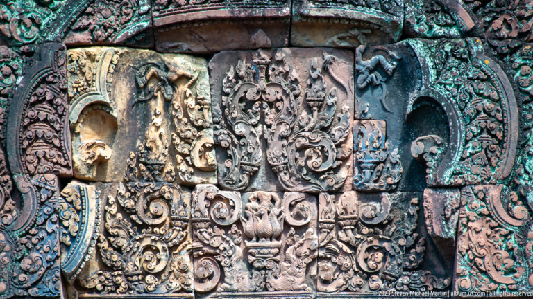 Close sandstone relief with small standing figures, a central face, and dense curling foliage.