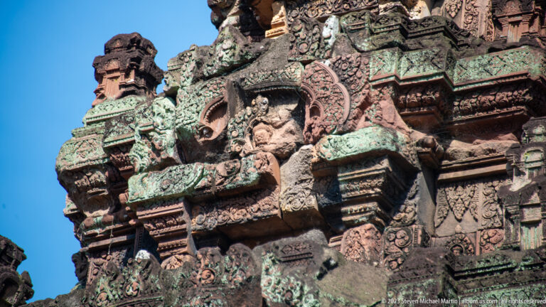 Upward view of carved tower projections, small figures, and green-stained sandstone against blue sky.
