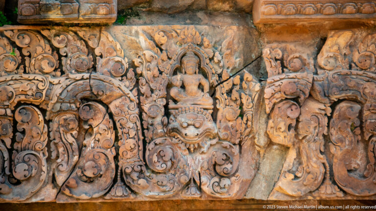 Rectangular sandstone lintel with a seated figure, central mask, and mirrored vegetal scrollwork.