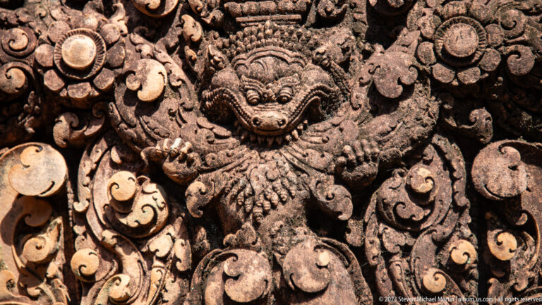 Extreme close-up of a fierce carved face with bulging eyes and fangs amid floral ornament.