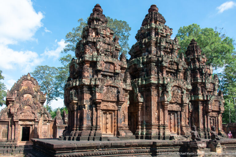 Three tall carved sandstone sanctuary towers on a shared platform beside a lower temple building.