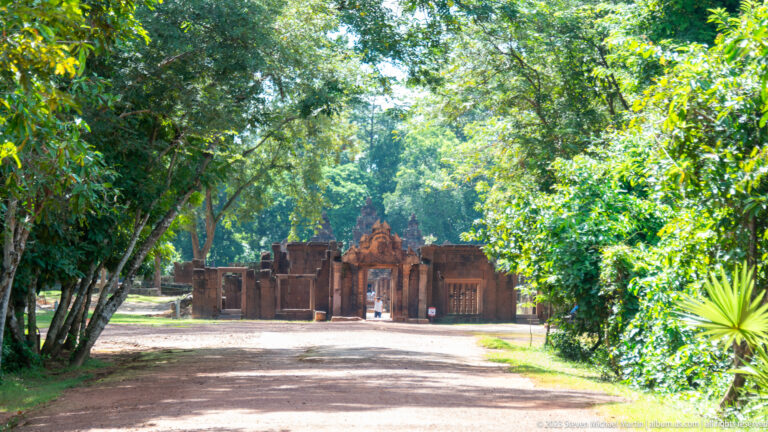 Tree-lined dirt approach leading to the distant pink sandstone entrance gateway of Banteay Srei.