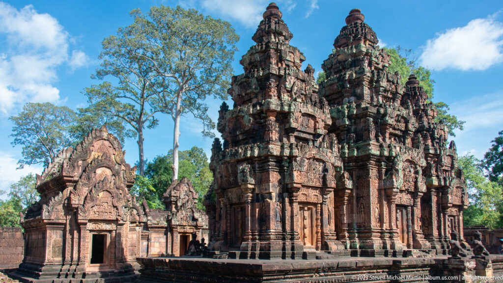 Sunlit view of three carved sandstone sanctuary towers beside a lower temple building and tall trees.
