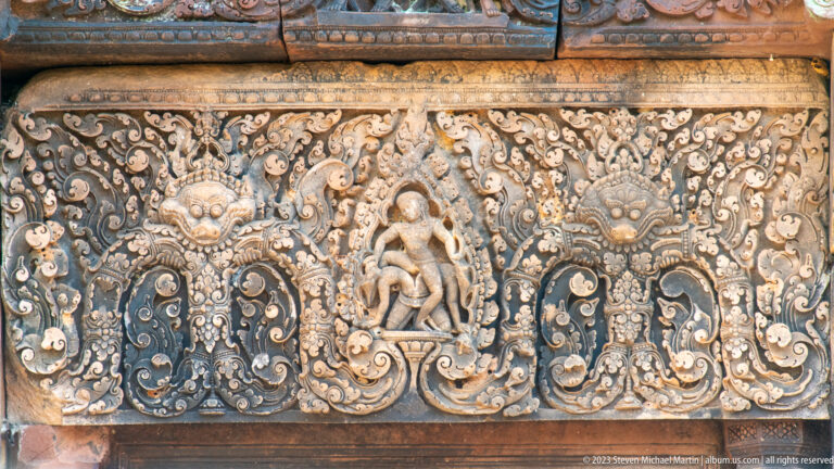 Rectangular sandstone lintel with a central female figure, two fierce masks, and mirrored floral scrolls.