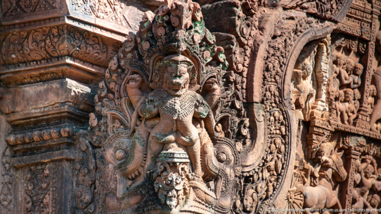 Standing crowned female figure in high relief on a sandstone corner beside a carved pediment.