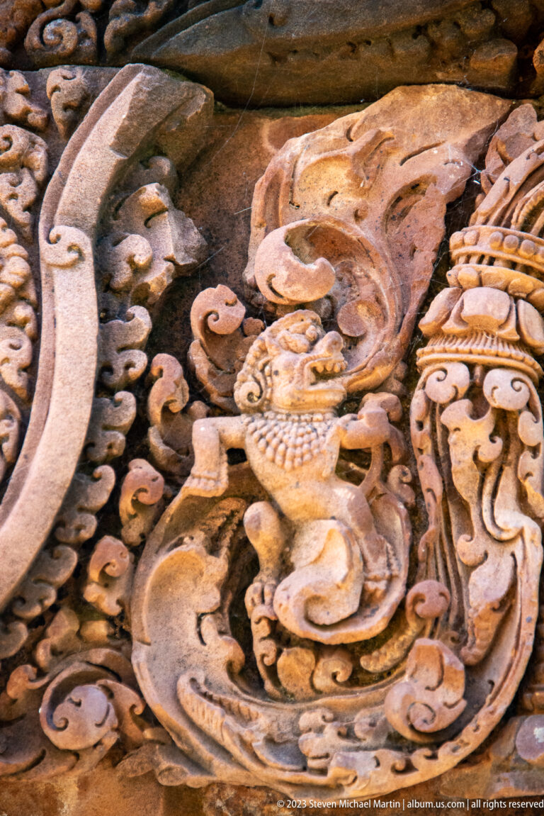 Profile of a small carved mythical bird with a crest and curling tail amid sandstone foliage.