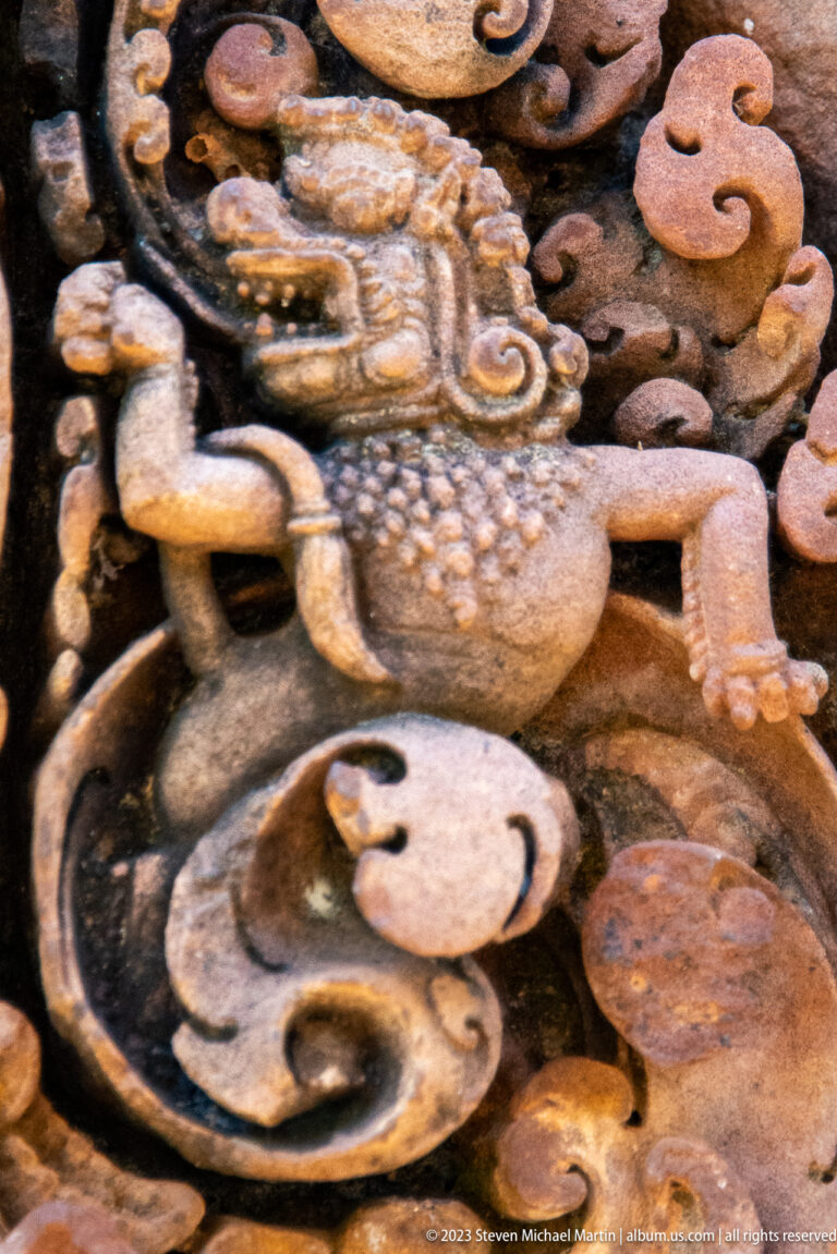 Close carved profile of a small lionlike mythical creature amid curling sandstone foliage.